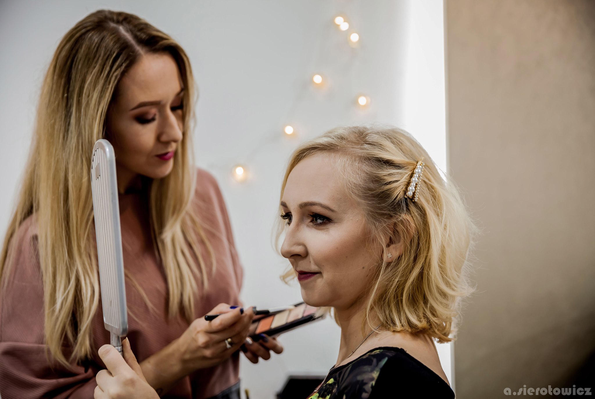 Wizażystka aplikuje makijaż pędzlem z palety na twarz klientki z blond włosami upiętymi spinką z pereł, oświetlenie w tle.
