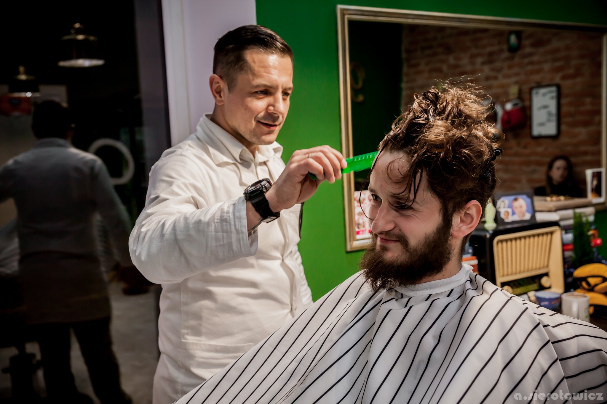 Fryzjer w Nowym Sączu czesze brodatego mężczyznę zielonym grzebieniem w salonie fryzjerskim z zieloną ścianą i lustrem.