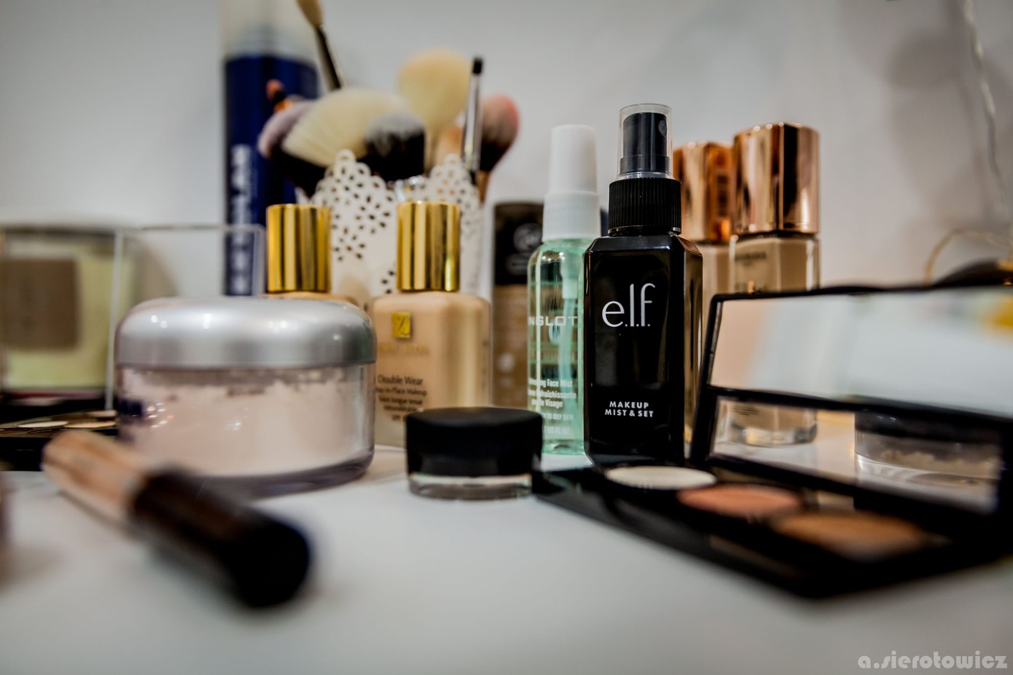 Zestaw kosmetyków do makijażu: puder, podkłady w odcieniach beżu i brązu, pędzle, mgiełka utrwalająca marki e.l.f. oraz paleta cieni do powiek, ułożone na białym blacie.