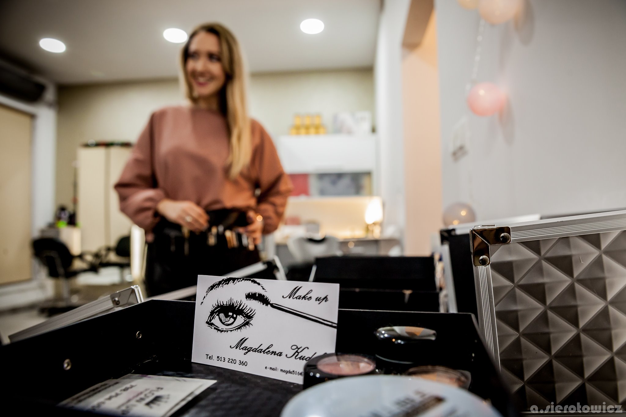 Wizażystka w tle, na pierwszym planie wizytówka z rysunkiem oka i napisem 'Make up Magdalena Kud' oraz kosmetyki w kuferku.