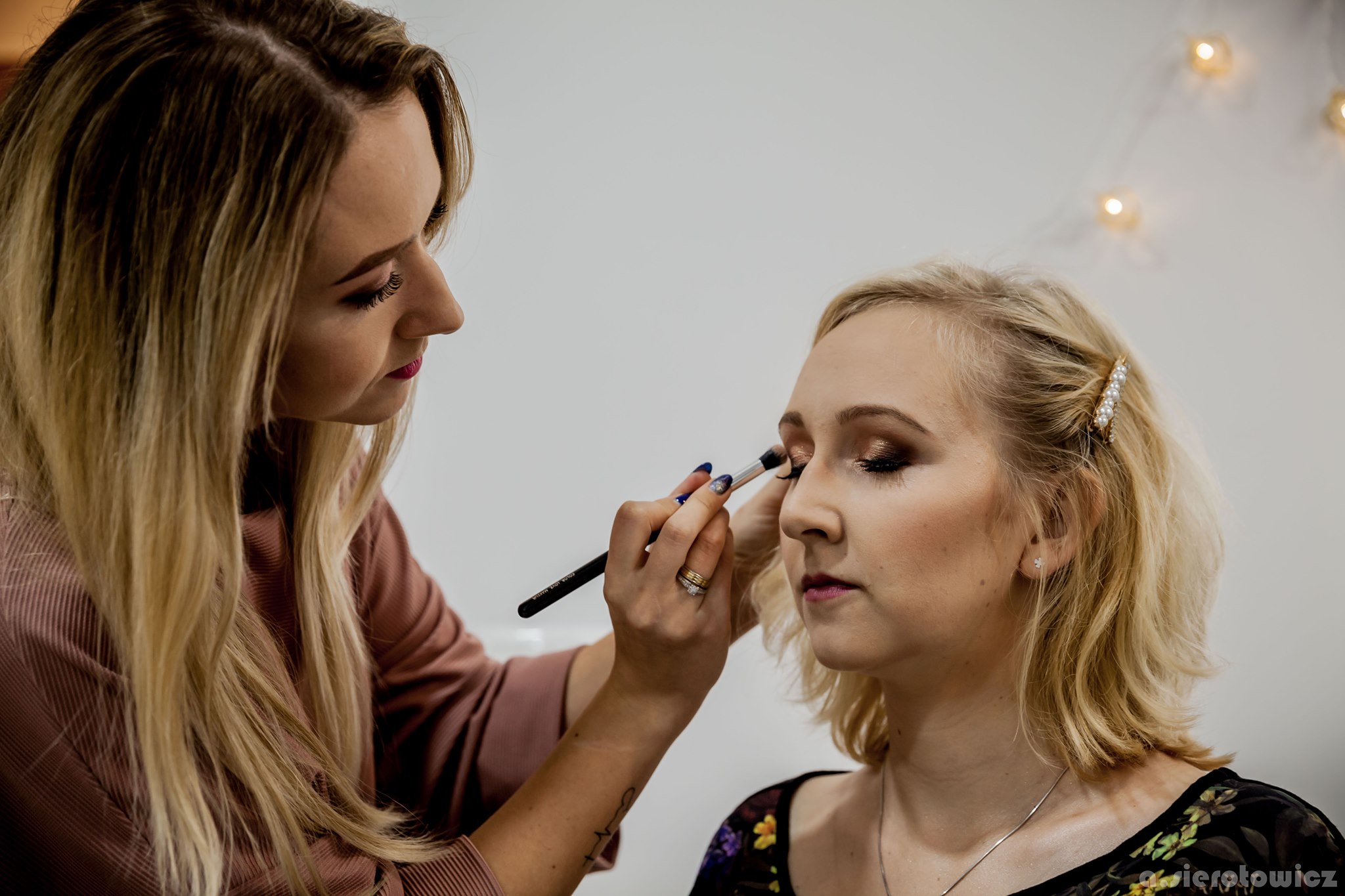 Makijażystka aplikuje cień do powiek pędzlem na powiekę klientki z blond włosami spiętymi ozdobną spinką z perełkami, w tle biała ściana z delikatnym oświetleniem.
