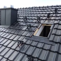 Nowy dach z ciemnej dachówki ceramicznej z zamontowanymi uchwytami do paneli słonecznych i kominem wentylacyjnym oraz oknem dachowym w trakcie montażu.