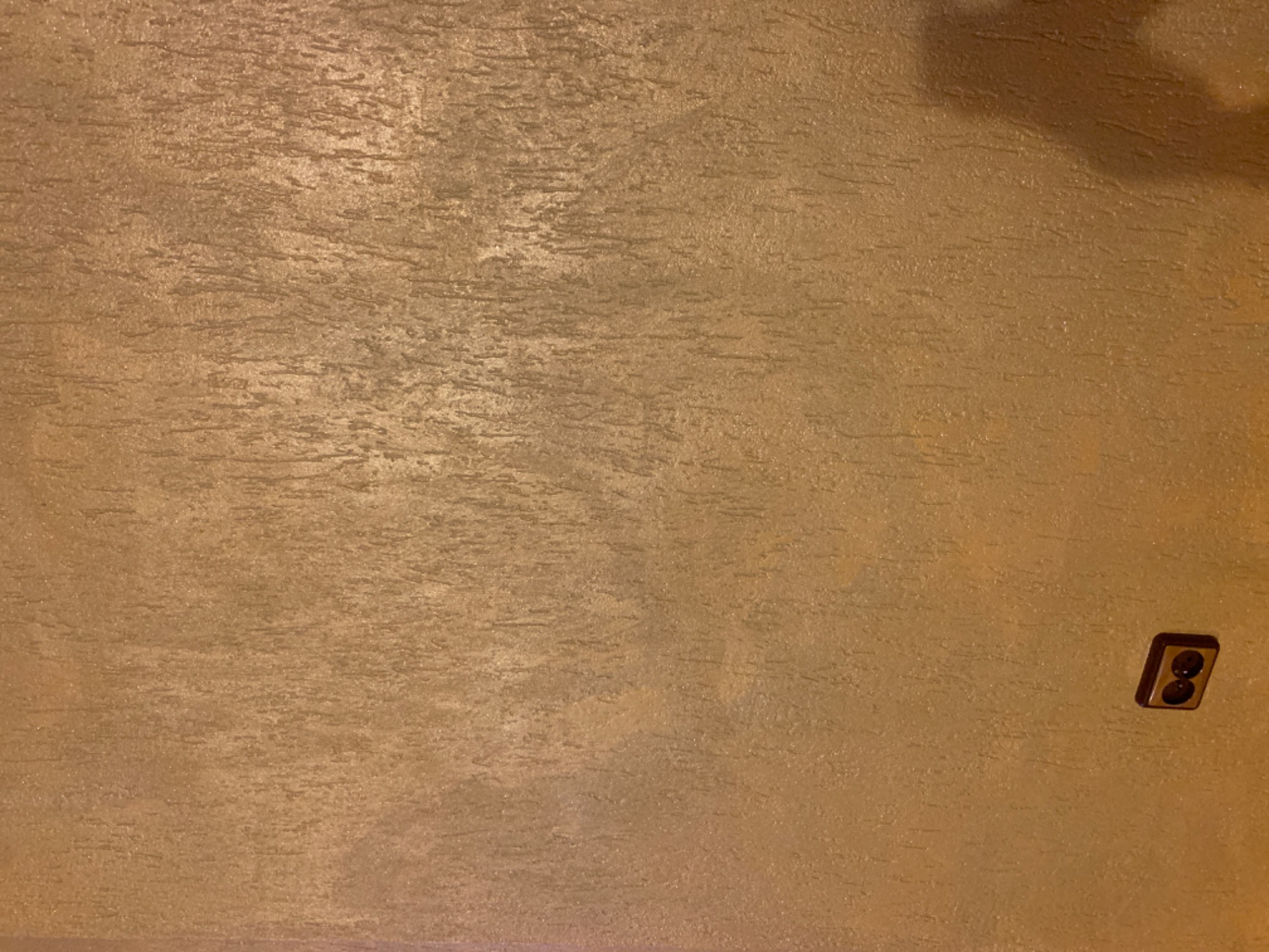 Teksturowana, brązowo-złota ściana wewnętrzna z gniazdkiem elektrycznym w prawym dolnym rogu.