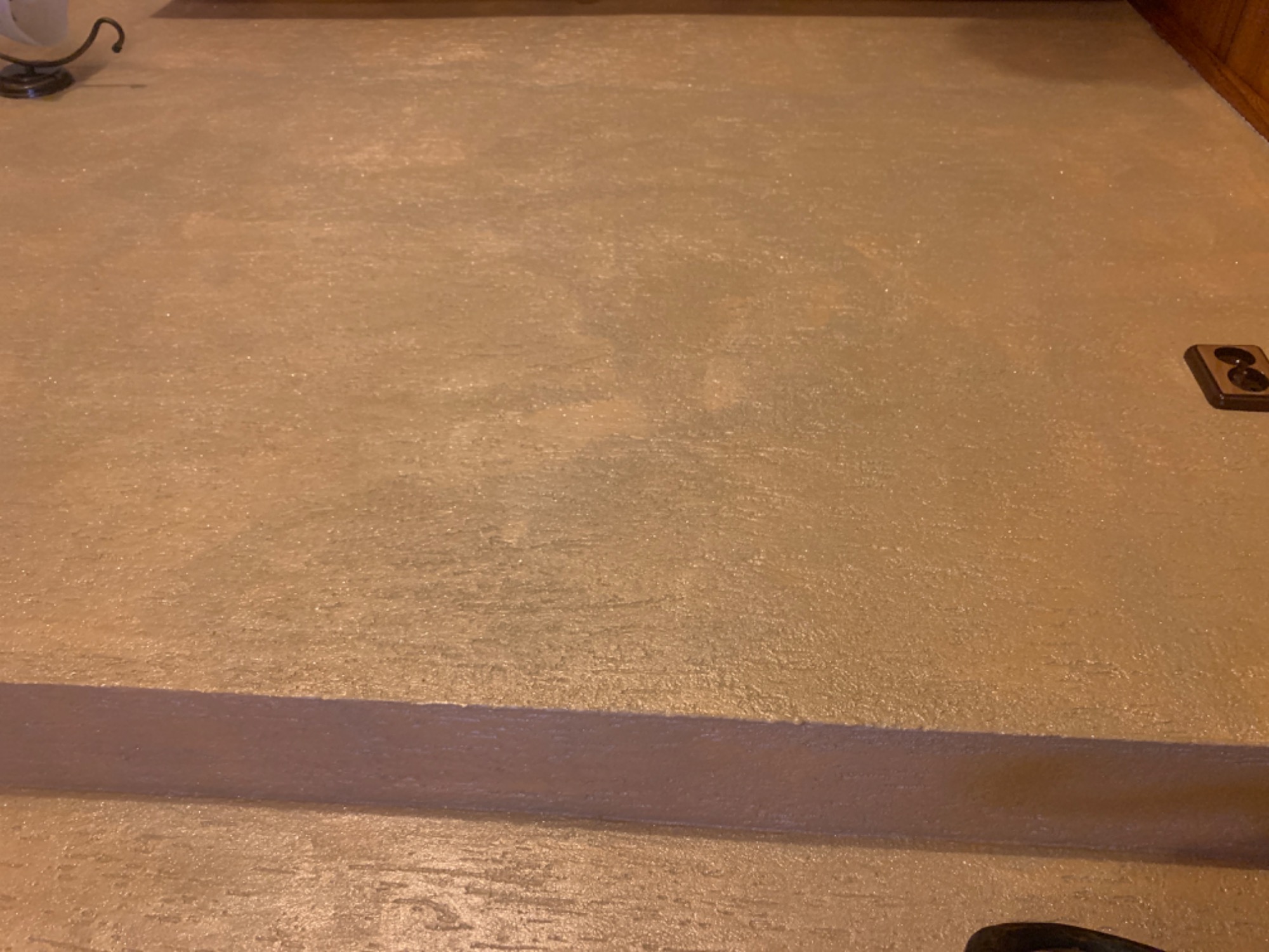 Beżowa, błyszcząca powierzchnia podłogi z widocznym progiem i gniazdkiem elektrycznym w rogu, oświetlona ciepłym światłem.