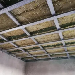 Montaż izolacji z wełny mineralnej na poddaszu, widoczne metalowe profile konstrukcyjne i białe sznurki podtrzymujące izolację przed zamontowaniem płyt gipsowo-kartonowych.