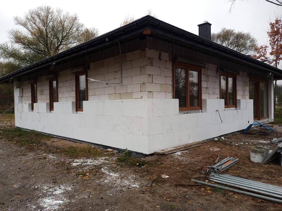 Dom jednorodzinny w trakcie ocieplania ścian zewnętrznych białym styropianem, widoczne okna z brązowymi ramami, teren wokół domu niezagospodarowany, widoczne narzędzia i materiały budowlane.