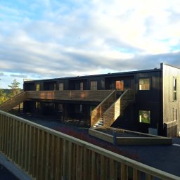 STOLBUD Wojciech Duda - Dwukondygnacyjny, czarny budynek szkieletowy z drewnianymi schodami zewnętrznymi i balustradami. Błękitne niebo z chmurami w tle. Widok z poziomu gruntu.