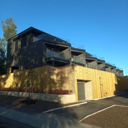 STOLBUD Wojciech Duda - Nowoczesny, trzypiętrowy dom szkieletowy z balkonami i drewnianym ogrodzeniem, widziany z dołu pod kątem. Błękitne niebo w tle.