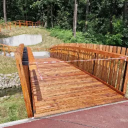 Nowy drewniany mostek z balustradami w parku, otoczony zielenią. Czerwono-biała taśma zabezpieczająca. Ujęcie z niskiej perspektywy.