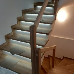 Klatka schodowa z jasnego drewna z podświetleniem LED na każdym stopniu, widoczne zabezpieczenia na stopniach, balustrada z prostych słupk&oacute;w.