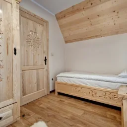 Jasne, drewniane wnętrze pokoju z łóżkiem, szafą i drzwiami, zdobione rzeźbieniami w motywy roślinne i geometryczne, poddasze wykończone boazerią.