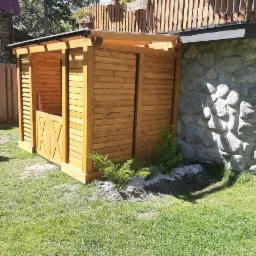 Drewniana konstrukcja altany z lamelami i dekoracyjnymi drzwiczkami na trawniku, przylegająca do kamiennej ściany budynku. Małe krzewy rosną u podstawy.