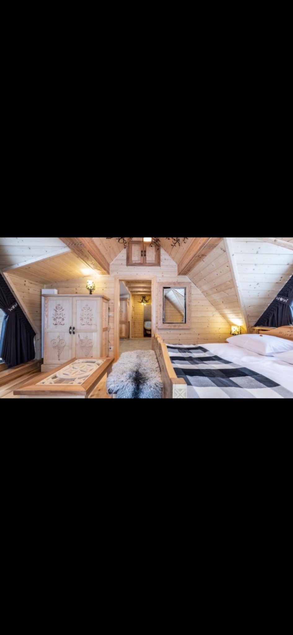 Drewniana sypialnia w góralskim stylu: boazeria, rzeźbiony kredens, łóżko z narzutą w kratę. Widok na łazienkę przez otwarte drzwi. Ciepłe oświetlenie.