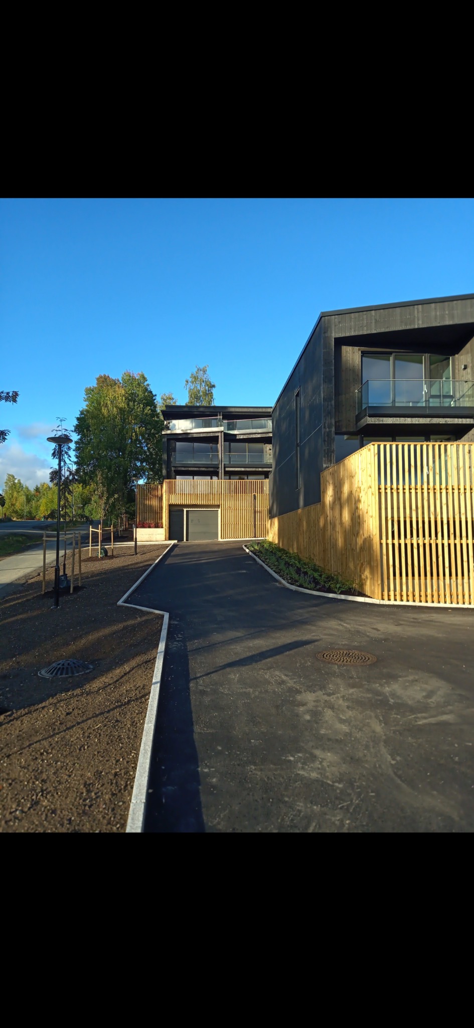 Nowoczesny dom szkieletowy z ciemną elewacją i jasnym drewnianym ogrodzeniem, widok z podjazdu w słoneczny dzień. Minimalistyczna architektura, proste linie, naturalne materiały.