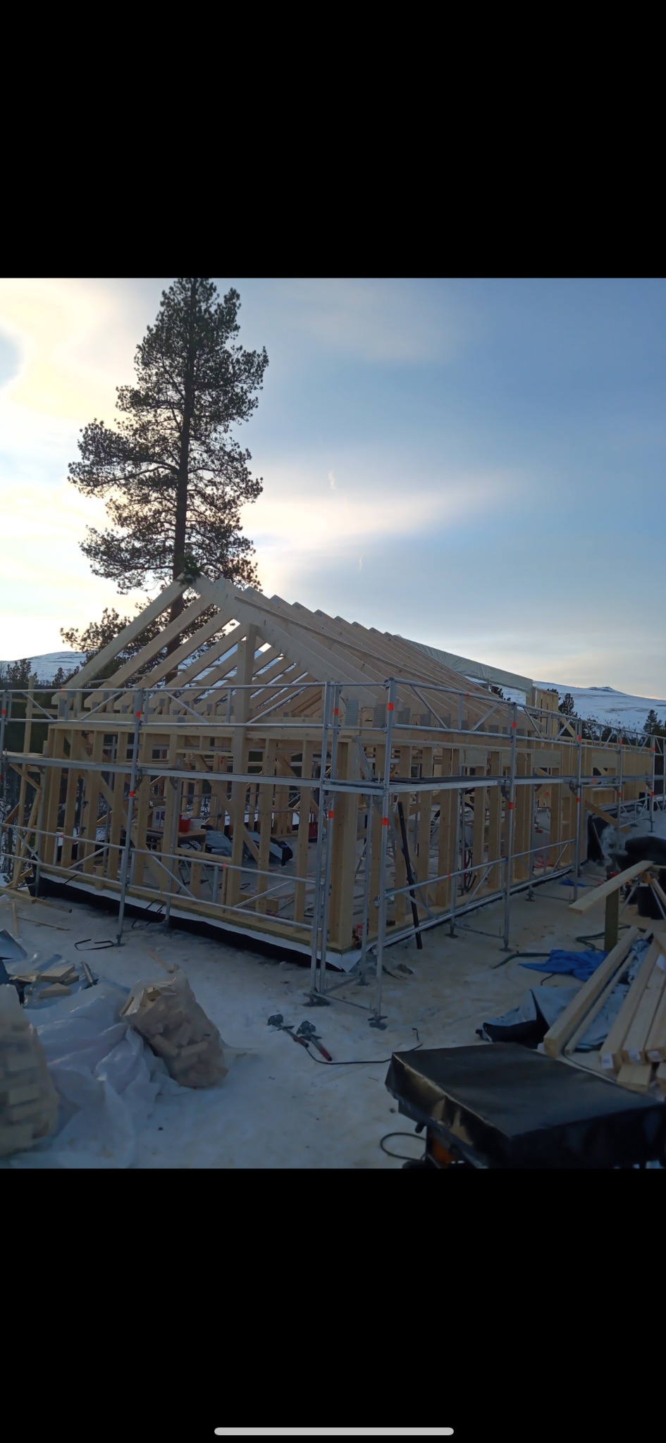 Wznoszenie szkieletu drewnianego domu w górach, z widocznymi rusztowaniami i narzędziami. W tle iglaste drzewo i ośnieżone szczyty, błękitne niebo.