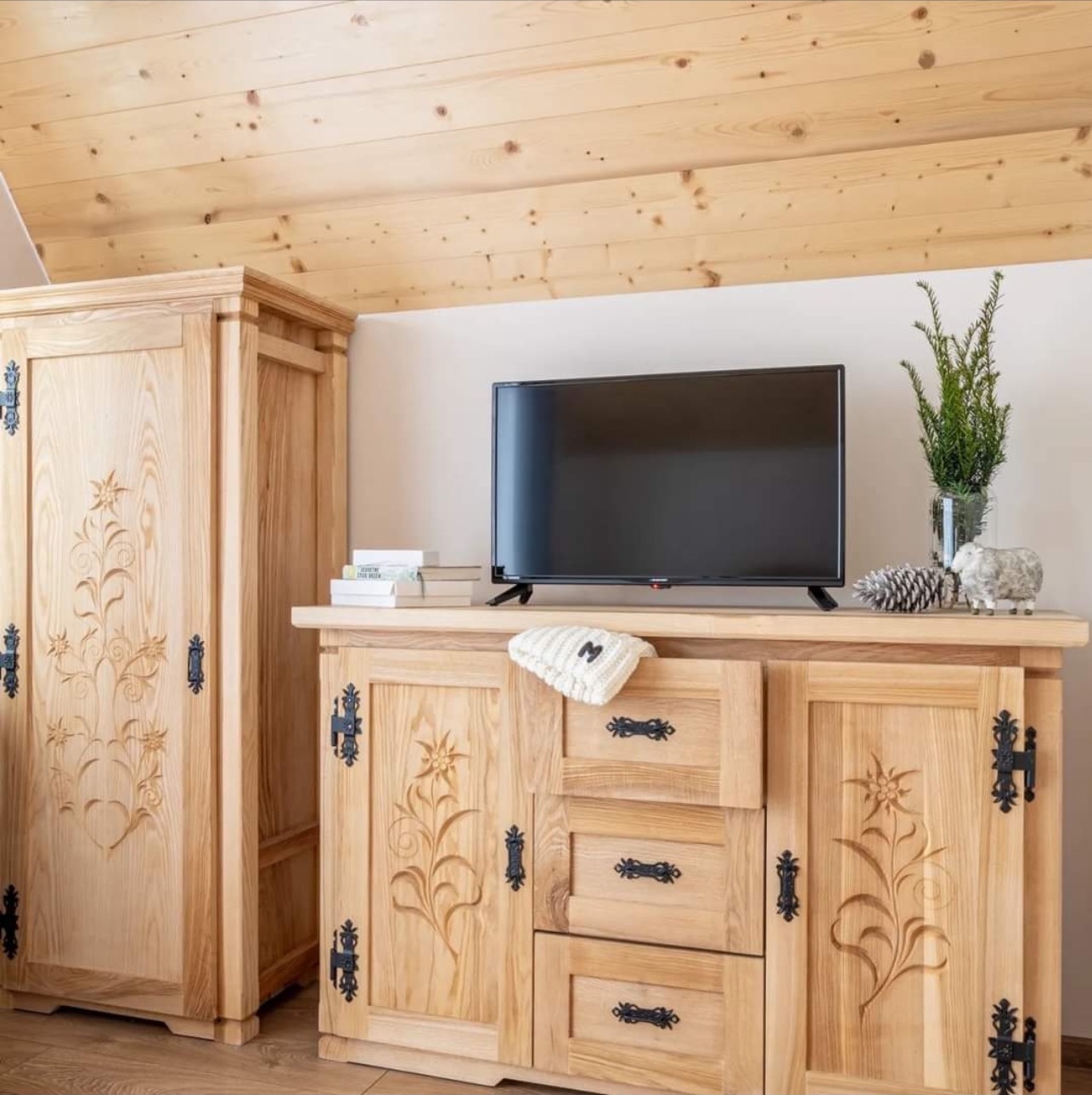 Drewniana szafa i komoda z rzeźbionymi motywami kwiatowymi i metalowymi okuciami w rustykalnym wnętrzu z drewnianym sufitem.