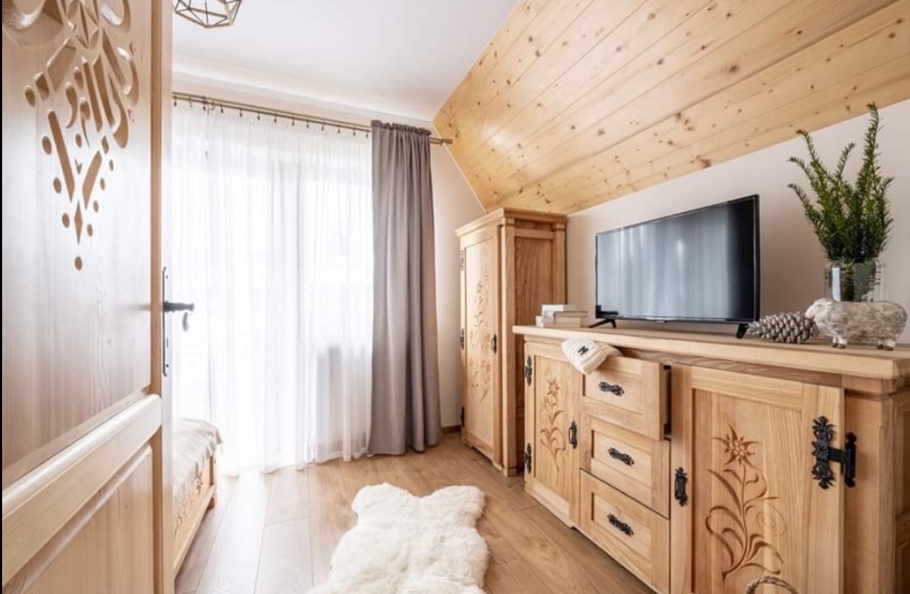 Jasne drewniane meble w góralskim stylu, zdobione rzeźbieniami, w pokoju z drewnianym sufitem i oknem z zasłonami. Otwarta, rzeźbiona drewniana drzwi.