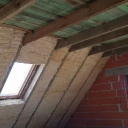 Poddasze w trakcie ocieplania wełną mineralną, widoczne drewniane belki stropowe, okno dachowe i murowana ściana z dwoma oknami.