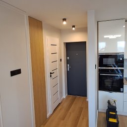 Dream House Ivan Vavryniuk - Nowoczesna kuchnia z białymi, lakierowanymi szafkami, marmurowym backsplashiem i drewnianym blatem, widoczny piekarnik i płyta indukcyjna, na podłodze panele, obok drzwi wejściowe, w rogu skrzynka...