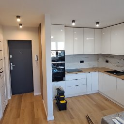 Dream House Ivan Vavryniuk - Nowoczesna, biała kuchnia z drewnianymi akcentami, czarnym zlewem i sprzętem AGD, widoczna skrzynka z narzędziami w rogu, częściowo zasłonięte meble folią ochronną.