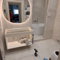 Dream House Ivan Vavryniuk - Montaż szafki pod umywalkę w łazience z podświetlanym lustrem, widoczne narzędzia i akcesoria glazurnicze na podłodze, płytki imitujące marmur na ścianach i podłodze, w tle wanna.