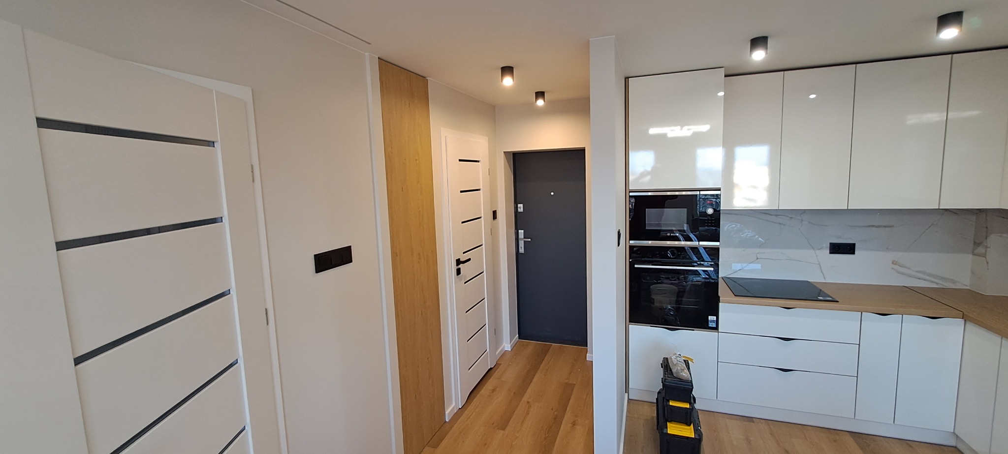 Nowoczesna kuchnia z białymi, lakierowanymi szafkami, marmurowym backsplashiem i drewnianym blatem, widoczny piekarnik i płyta indukcyjna, na podłodze panele, obok drzwi wejściowe, w rogu skrzynka...