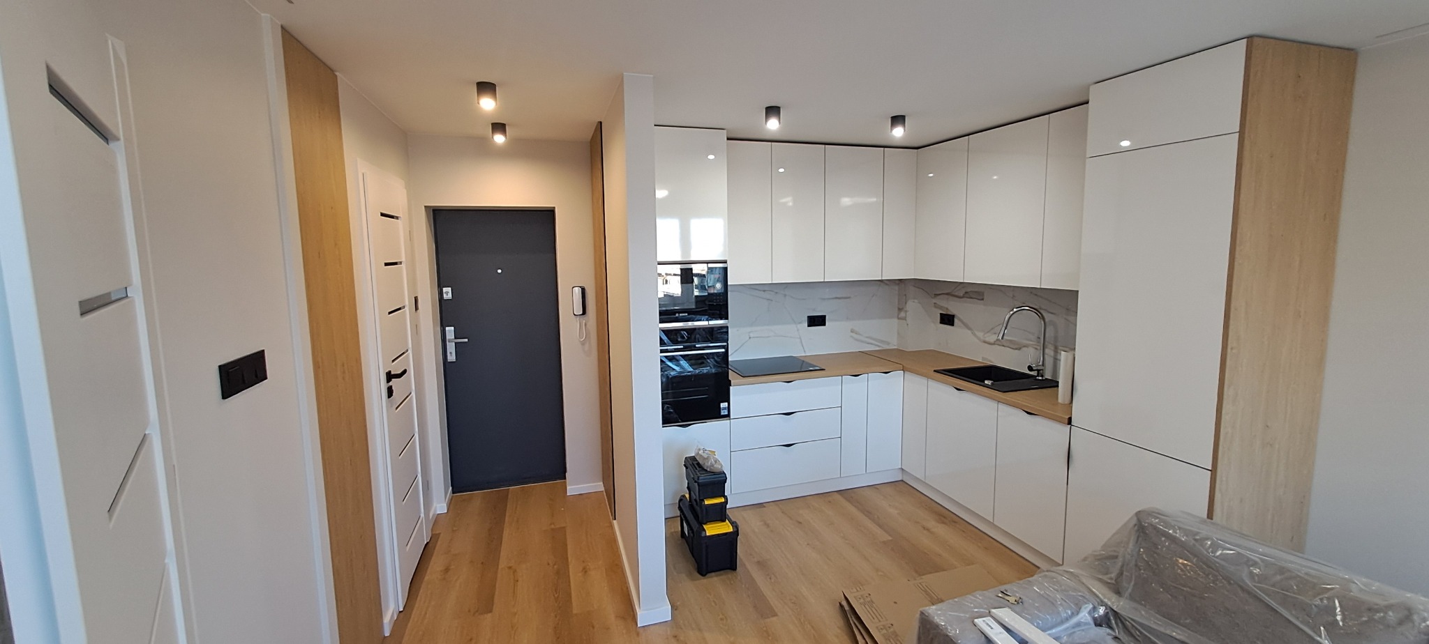 Nowoczesna, biała kuchnia z drewnianymi akcentami, czarnym zlewem i sprzętem AGD, widoczna skrzynka z narzędziami w rogu, częściowo zasłonięte meble folią ochronną.