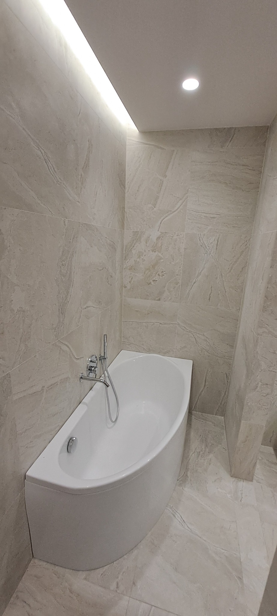 Nowoczesna łazienka z białą, asymetryczną wanną, chromowaną armaturą i beżowymi płytkami imitującymi marmur, oświetlona listwą LED podsufitową.