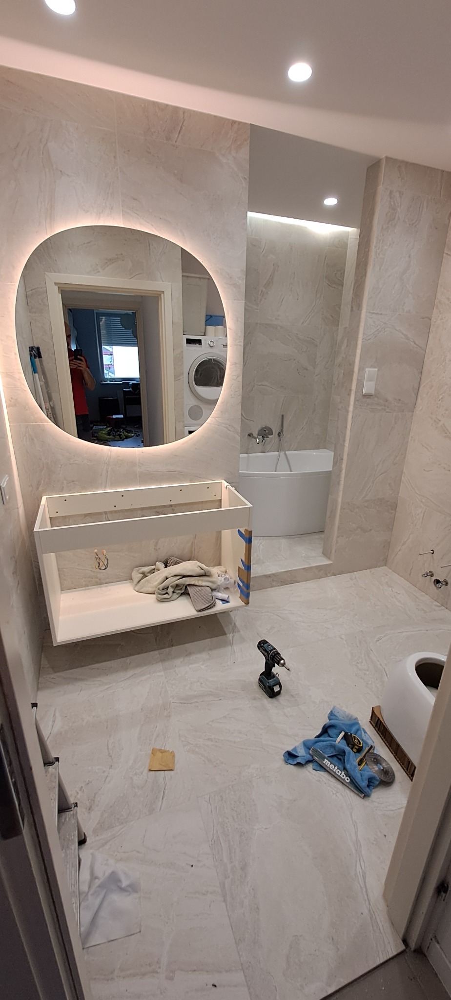 Montaż szafki pod umywalkę w łazience z podświetlanym lustrem, widoczne narzędzia i akcesoria glazurnicze na podłodze, płytki imitujące marmur na ścianach i podłodze, w tle wanna.