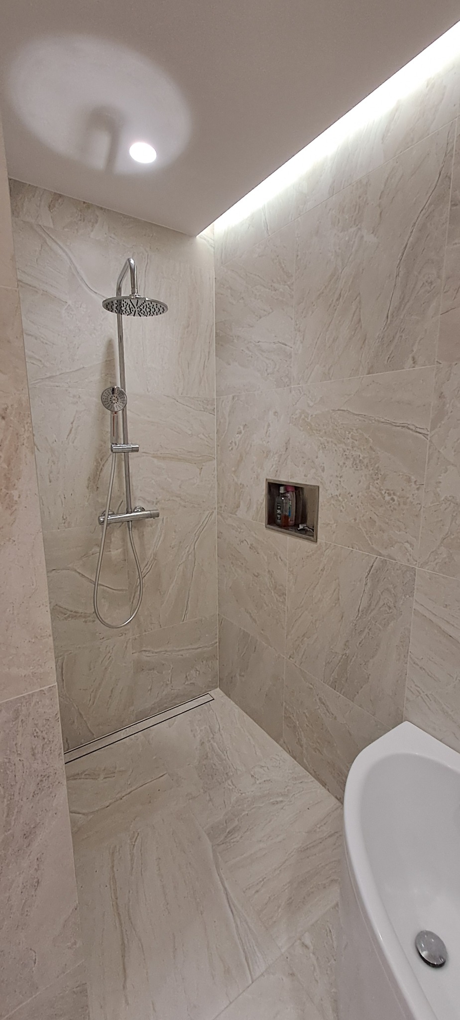 Nowoczesna kabina prysznicowa z deszczownicą i odpływem liniowym, wykończona beżowymi płytkami imitującymi marmur, z wnęką na kosmetyki i subtelnym oświetleniem LED.