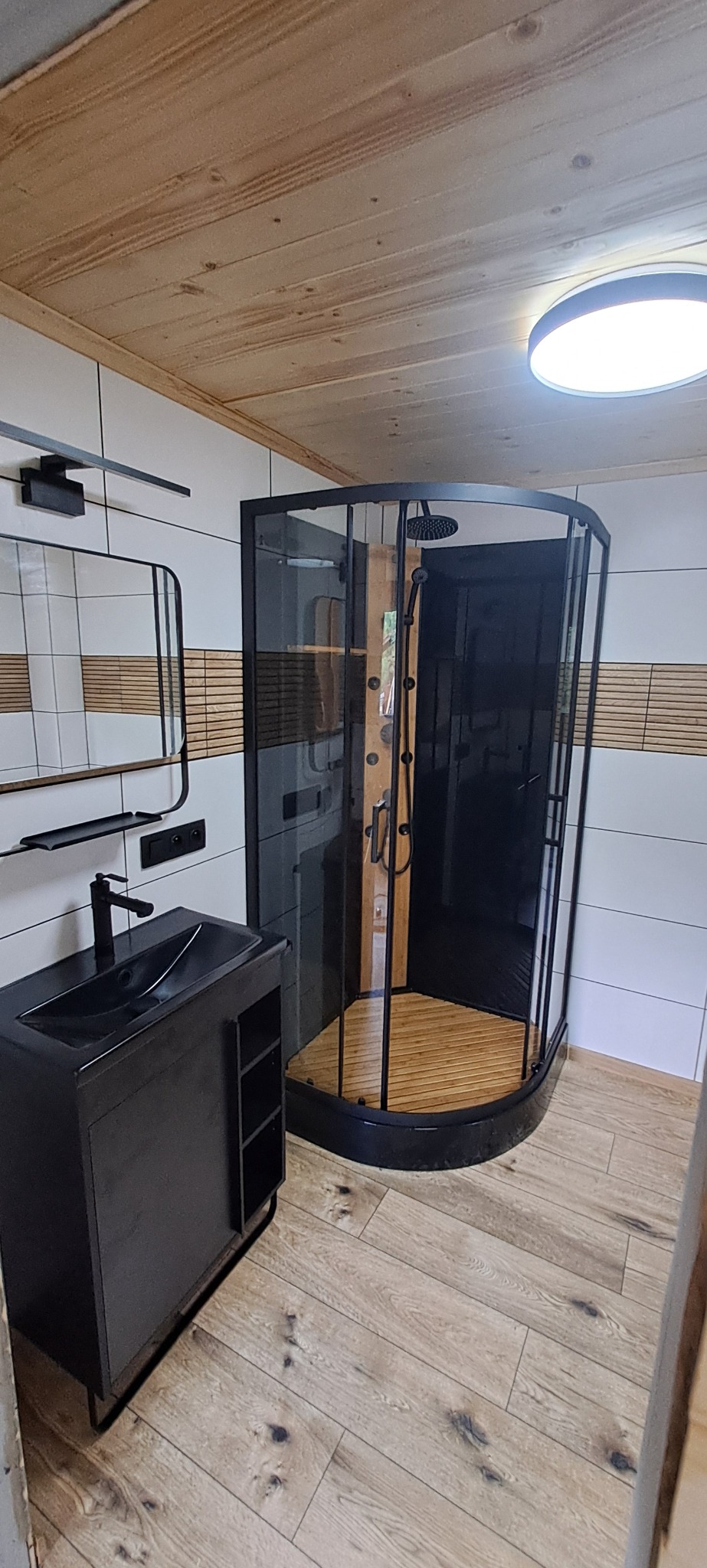 Nowoczesna łazienka z kabiną prysznicową typu walk-in z czarnymi ramami, czarną szafką pod umywalkę z półkami i lustrem z czarną ramą, białe płytki z dekoracyjnym pasem z drewnianych paneli...