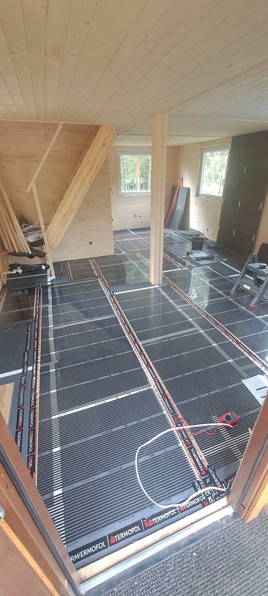 Montaż folii grzewczej pod podłogę w drewnianym domu, widoczne panele podłogowe i schody w tle.