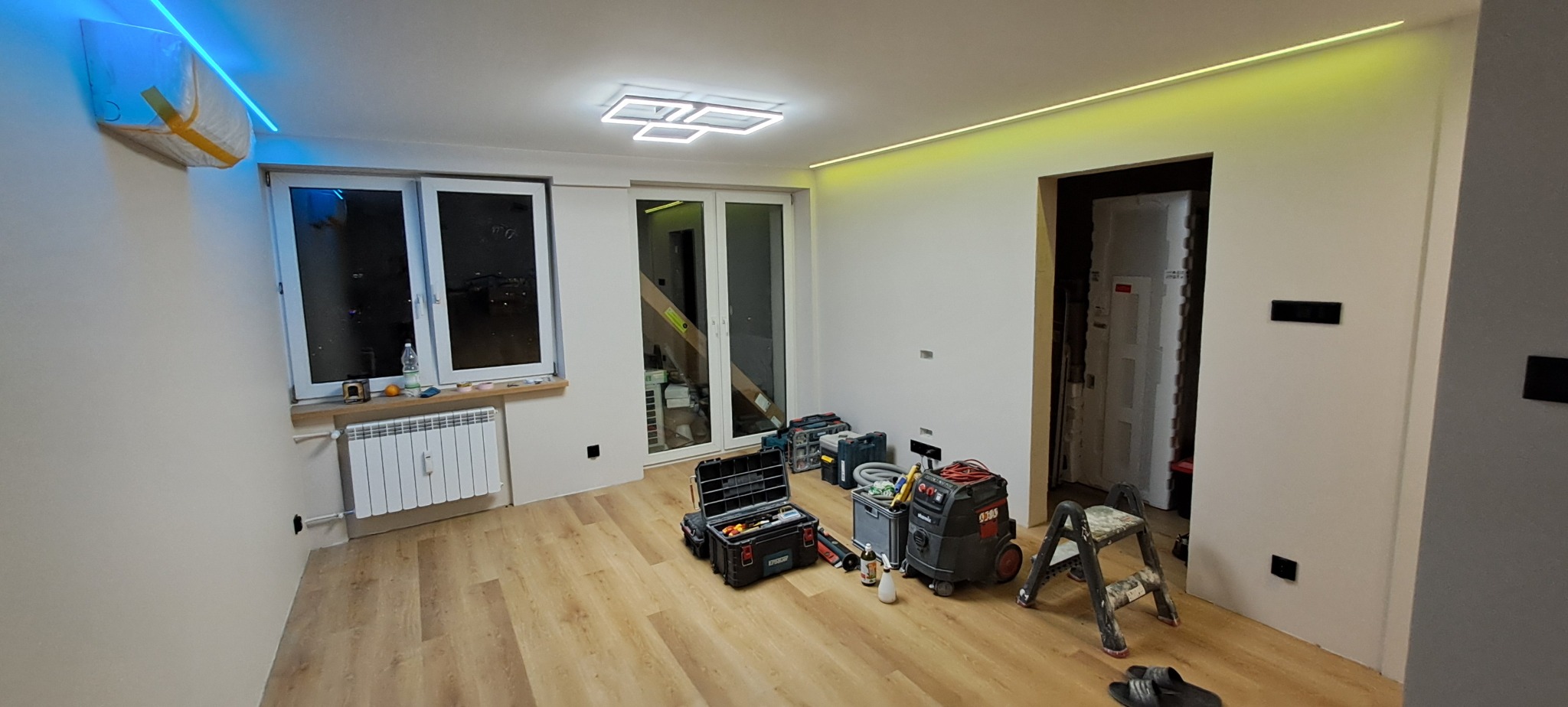 Wnętrze pokoju w trakcie prac wykończeniowych, widoczne narzędzia, drabina, odkurzacz przemysłowy, skrzynki z narzędziami, oświetlenie LED na suficie i pod sufitem, zabezpieczony klimatyzator folią...