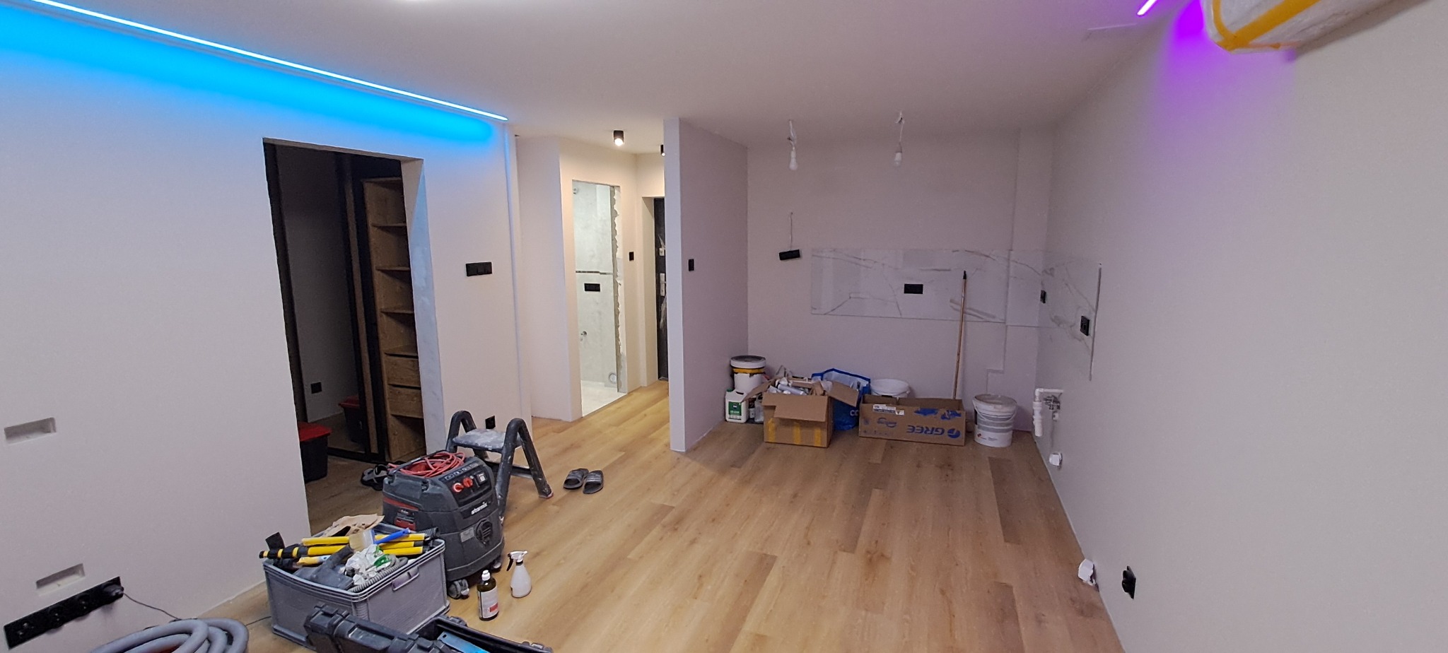 Wnętrze mieszkania w trakcie prac wykończeniowych; widoczne narzędzia, drabina, podłoga laminowana, przygotowane ściany, oświetlenie LED na suficie i częściowo ułożona glazura na ścianie w tle.