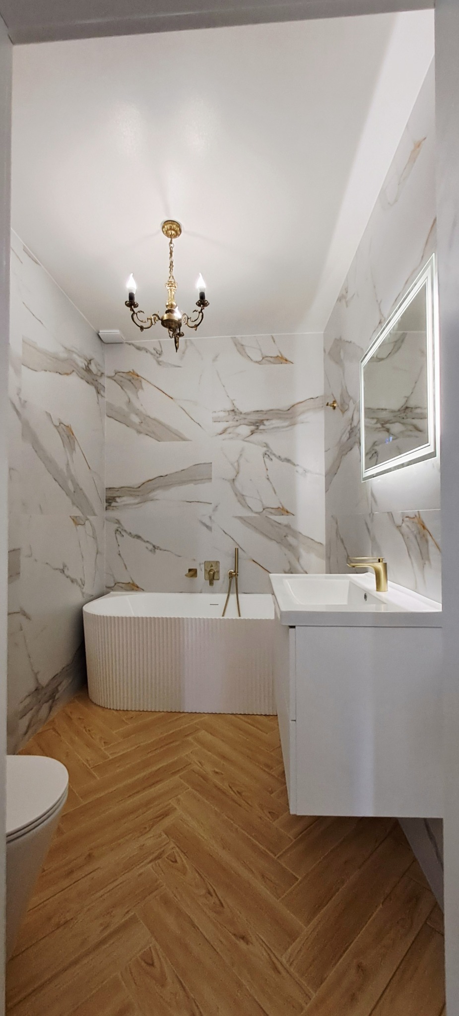 Elegancka łazienka po remoncie: marmurowe płytki, złota armatura, drewniana podłoga w jodełkę i designerski żyrandol.