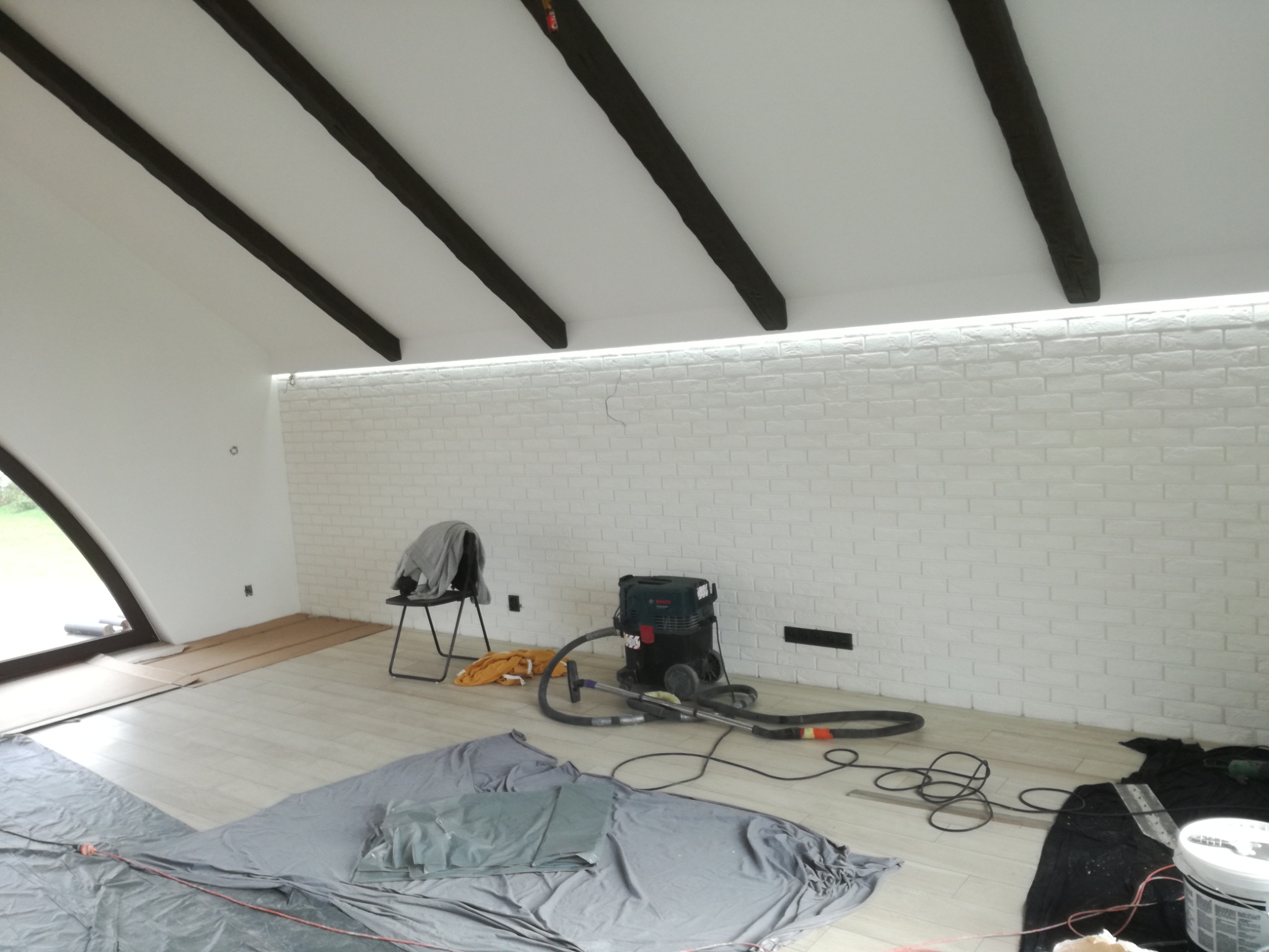 Skośny sufit z ciemnymi belkami i oświetleniem LED nad białą ścianą z cegły, podłoga zabezpieczona folią malarską, widoczny odkurzacz przemysłowy i krzesło z ubraniem.