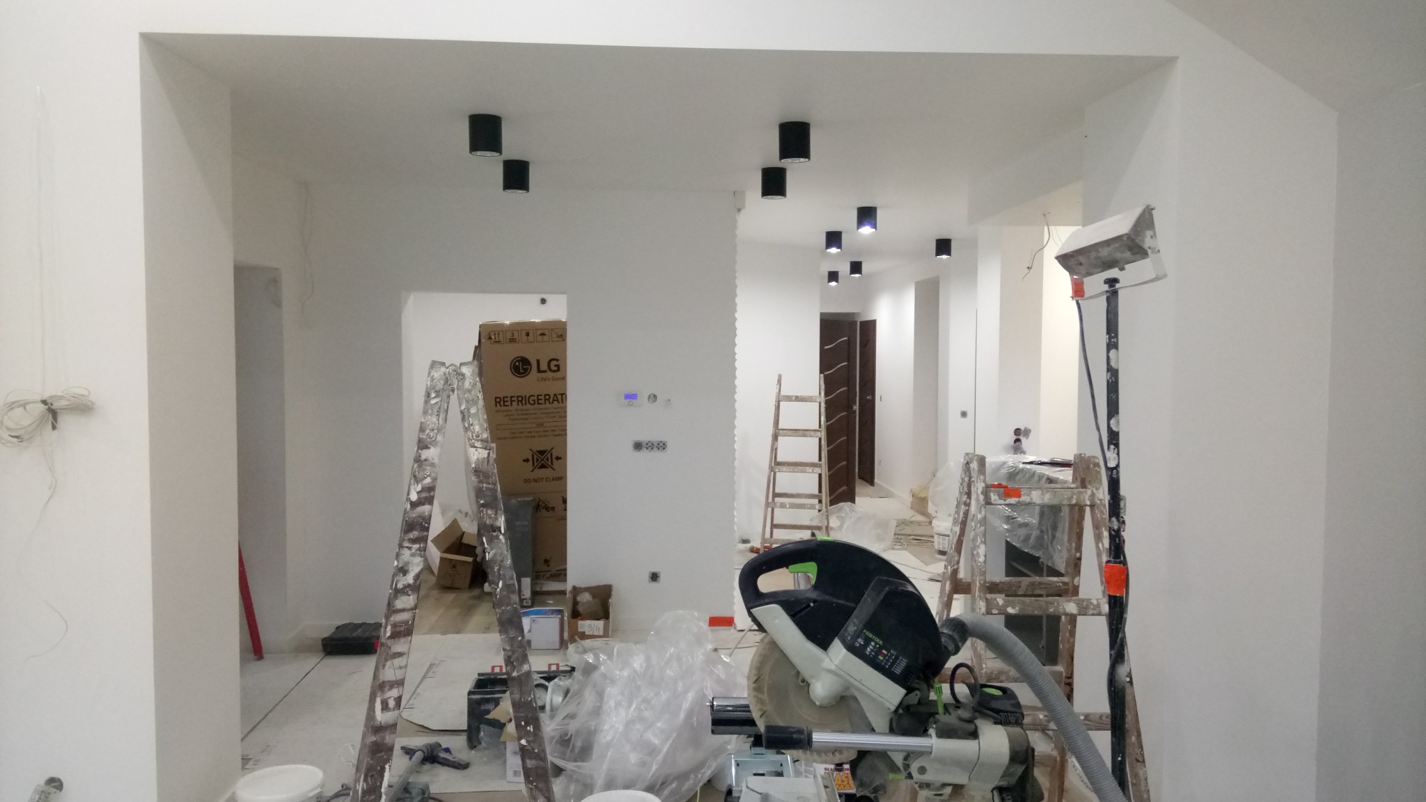 Wnętrze w trakcie prac wykończeniowych: widoczne ściany po gładziowaniu, czarne lampy sufitowe, drabiny i narzędzia, w tle karton po lodówce LG.