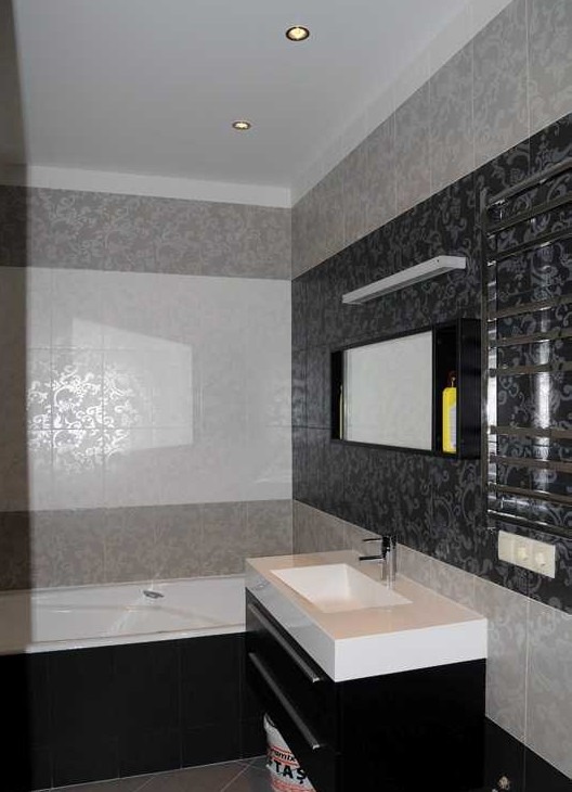 Nowoczesna łazienka z czarno-białymi płytkami w kwiatowe wzory, prostokątną umywalką nablatową na czarnej szafce z szufladami, grzejnikiem drabinkowym i ukrytą półką z oświetleniem.
