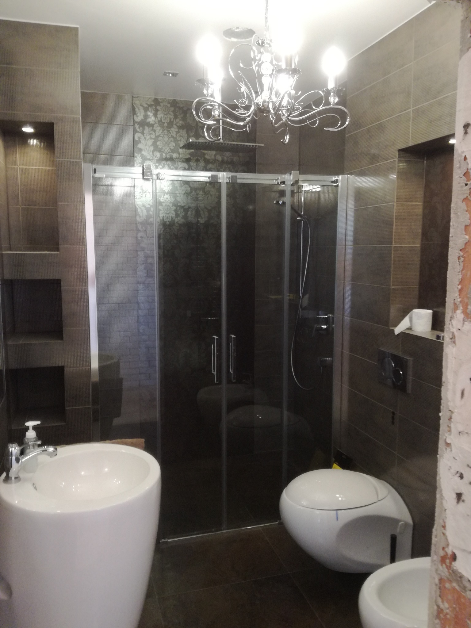 Nowoczesna łazienka z kabiną prysznicową ze szklanymi drzwiami przesuwnymi, umywalką na postumencie i podwieszaną toaletą, oświetlona ozdobnym żyrandolem, widoczny fragment niedokończonej ściany.