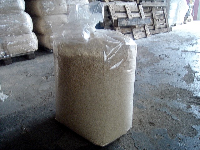 Worek z trocinami w magazynie, widoczne palety i inne worki w tle, podłoga z plamami.