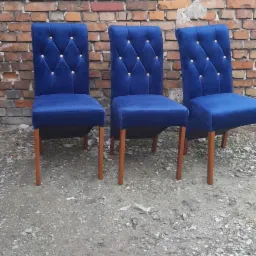Trzy odnowione krzesła z ciemnoniebieską tapicerką i złotymi guzikami, ustawione przed ceglaną ścianą na żwirowym podłożu.