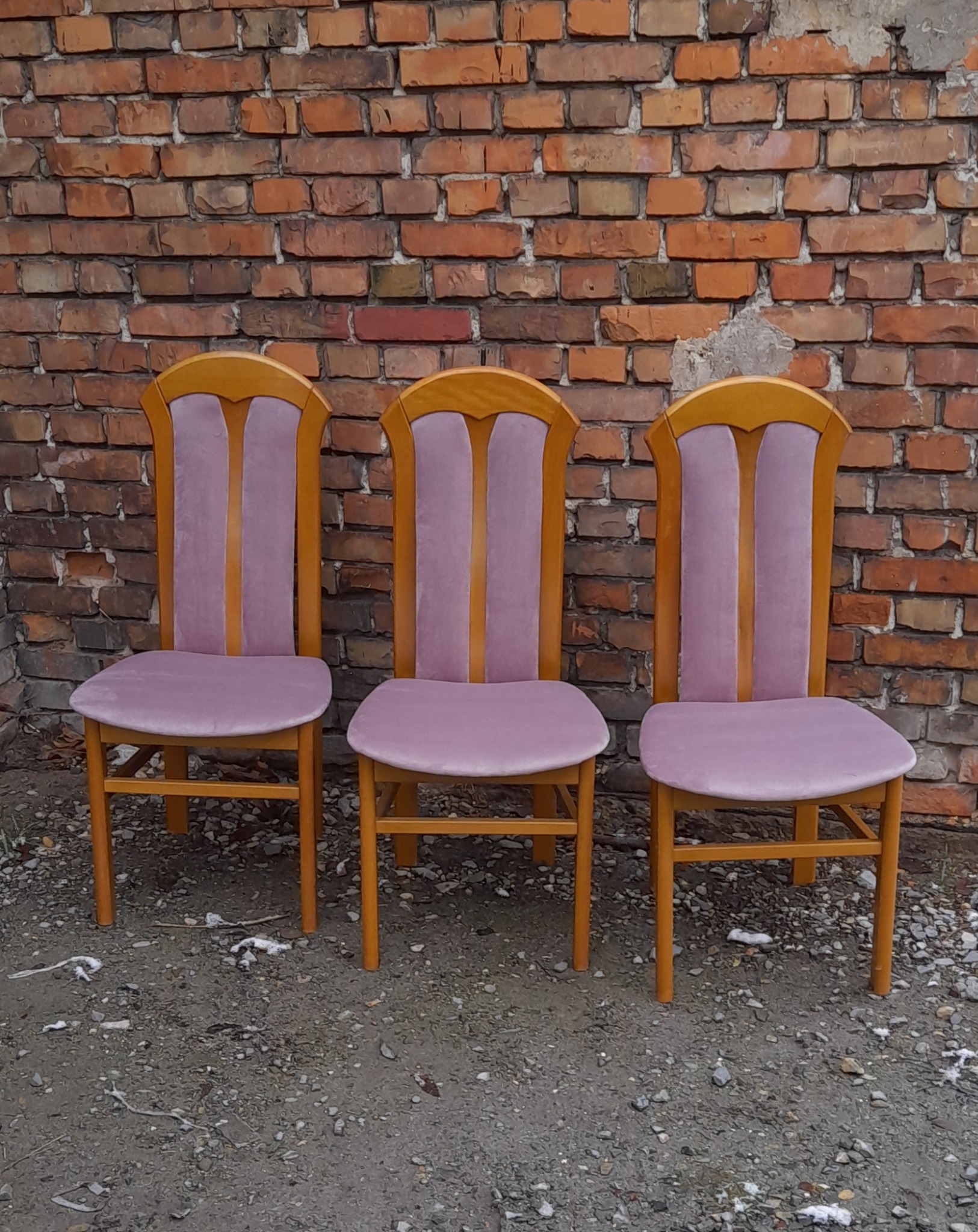 Trzy odnowione krzesła z jasnego drewna i fioletową tapicerką, ustawione na żwirowym podłożu przed czerwoną ceglaną ścianą z widocznymi śladami napraw.
