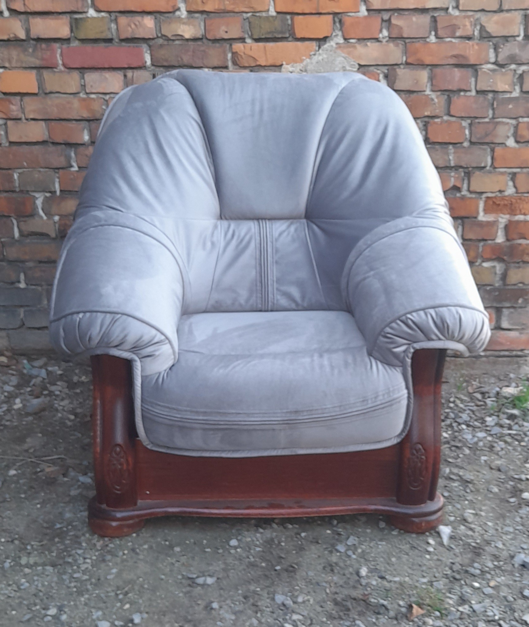 Szary, tapicerowany fotel z drewnianymi nogami i zdobieniami, ustawiony na żwirowym podłożu na tle ceglanej ściany.