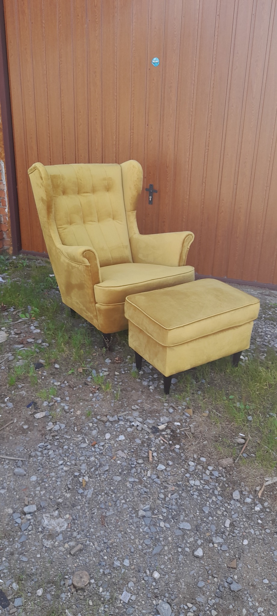Odrestaurowany fotel typu uszak i podnóżek w odcieniach żółci, ustawione na żwirowym podłożu przed brązową bramą garażową.