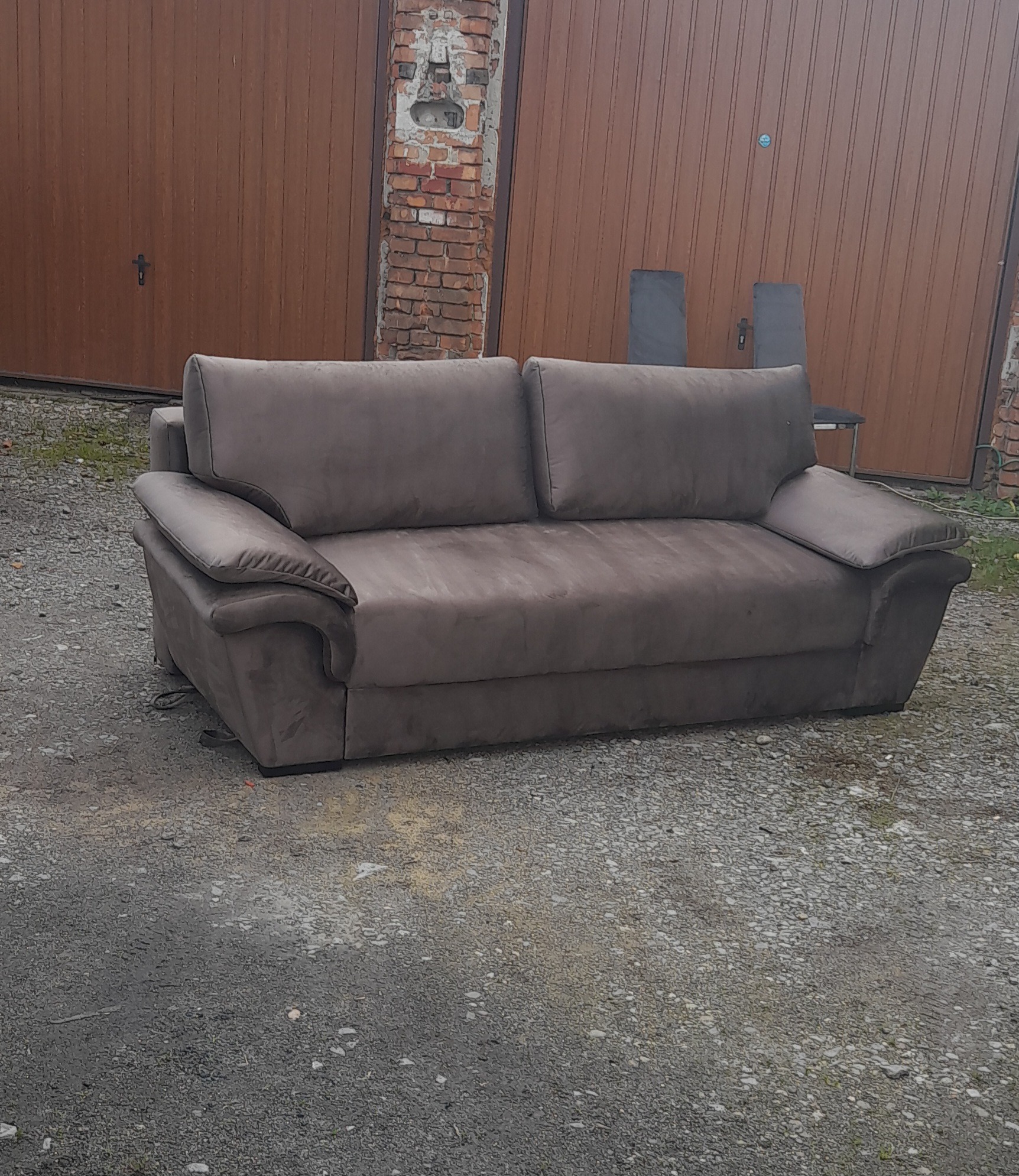 Szara sofa z poduszkami na zewnątrz, na żwirowym podłożu, z drewnianymi drzwiami garażowymi i fragmentem ceglanego muru w tle.