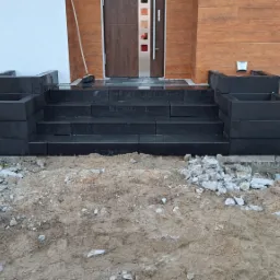 Nowe, czarne schody z kostki brukowej prowadzące do drzwi wejściowych domu, otoczone ziemią i gruzem przed zagospodarowaniem terenu.