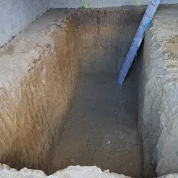 Głęboki wykop w ziemi z drabinką opartą o ścianę, widoczne bloczki betonowe i cegły na górze, zielone oznaczenia na ścianie wykopu.