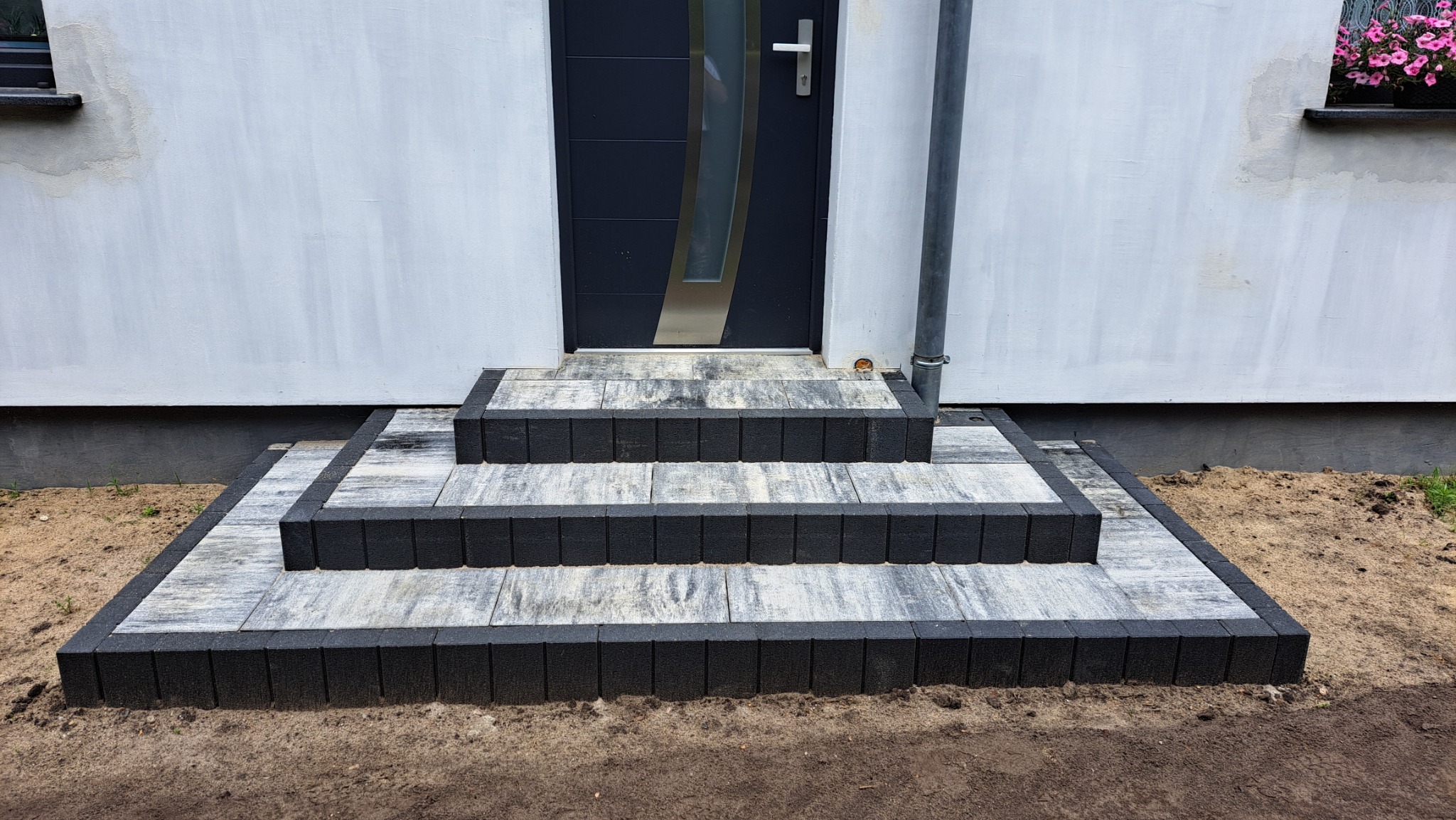 Nowe, trzy-stopniowe schody wejściowe z szaro-białej kostki brukowej z czarnym obramowaniem, prowadzące do nowoczesnych, grafitowych drzwi z metalową klamką, na tle białej elewacji domu z rynną...