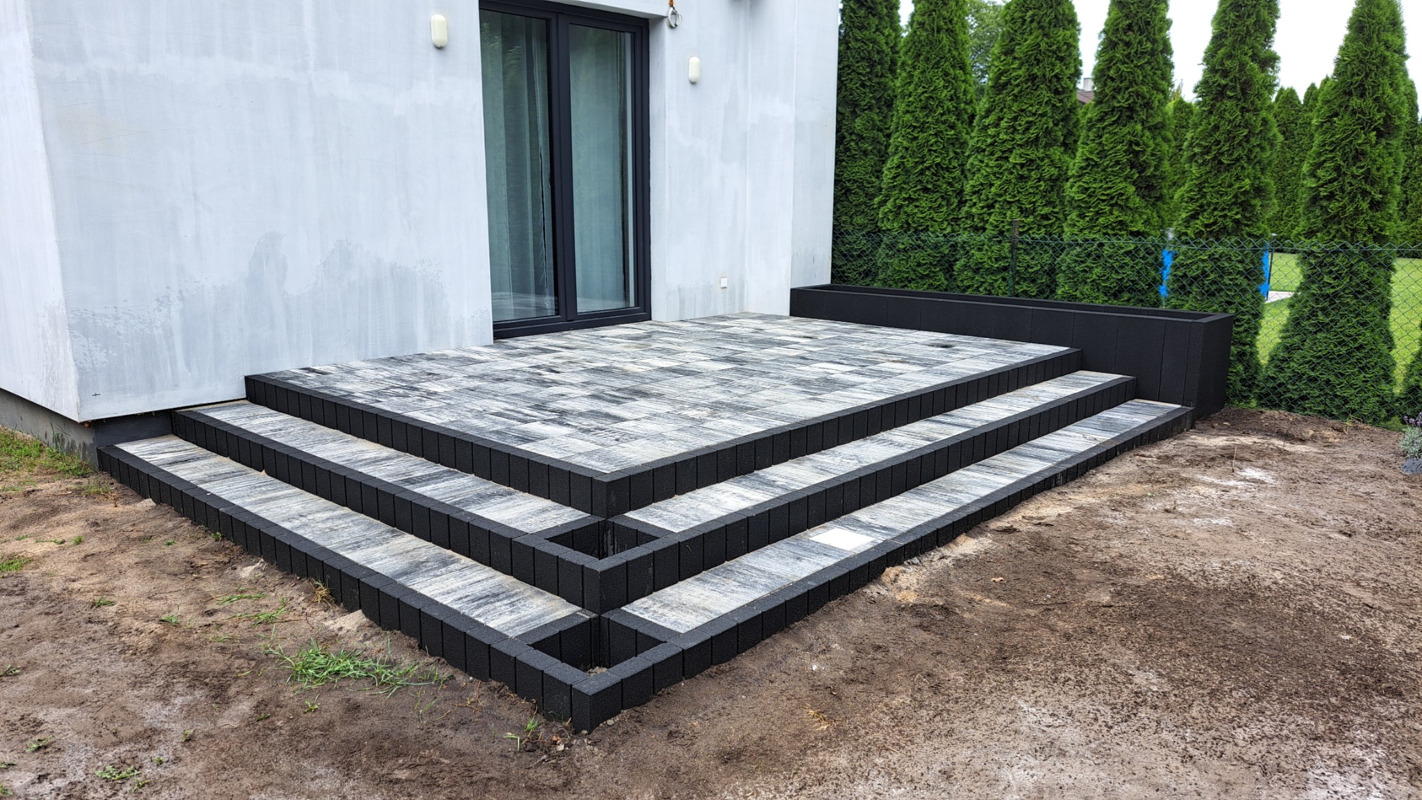 Schody z kostki brukowej prowadzące do drzwi tarasowych nowoczesnego domu, z czarnymi obrzeżami i szaro-białym wzorem kostki, na tle zielonych tuj i częściowo przygotowanego terenu.