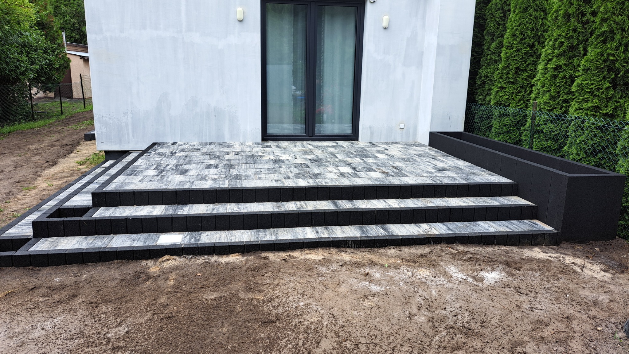 Nowoczesny taras z kostki brukowej w odcieniach szarości i czerni, prowadzący do domu w Strzałkowie. Widoczne trzy stopnie i donica w kolorze czarnym. Tło stanowi zielona roślinność i fragment...